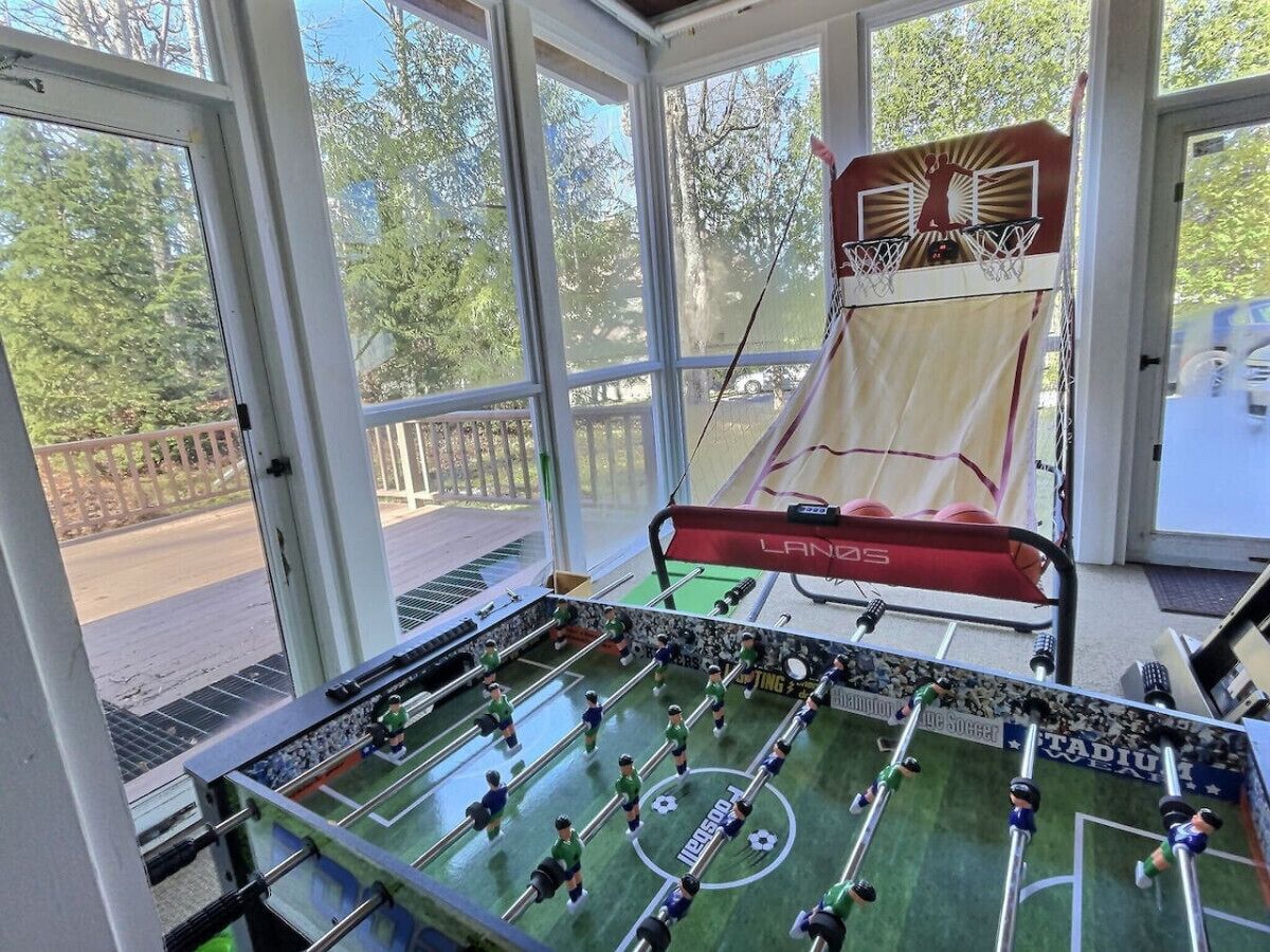Ein Tischkicker in einem Raum mit großen Fenstern, einem Basketballkorb und Blick auf Bäume und eine Straße.