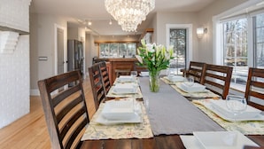 A dining area with a chandelier, wooden chairs, a long table set for six, and a view of the outdoors through large windows. - Bearities - 2 homes, sleeps 28, hot tubs, grills! (Killington)