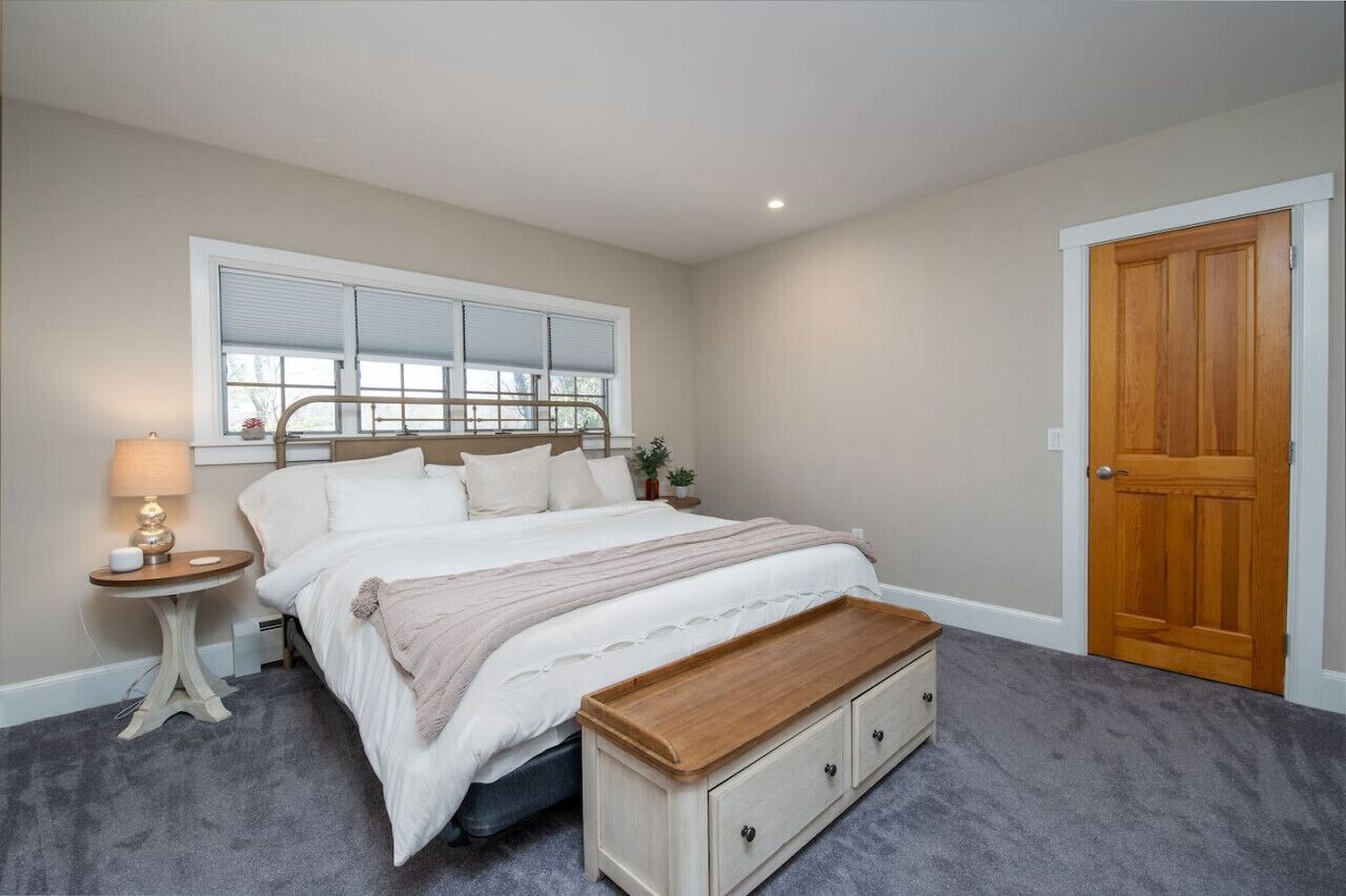 A bedroom with a bed, bedside table, lamp, window, and a wooden door.