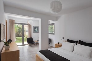 A modern bedroom with a large bed, a desk, and a chair. There are two windows providing natural light and a view of greenery outside.