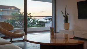 Ein modernes Wohnzimmer mit einem runden Holztisch, einem Korbstuhl, einem Blumentopf und Blick auf den Sonnenuntergang durch eine große Schiebeglastür.