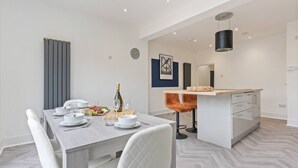 Un espace repas moderne avec une table en bois dressée pour quatre personnes, des chaises blanches et un coin bar agrémenté d’un tabouret de bar en cuivre.