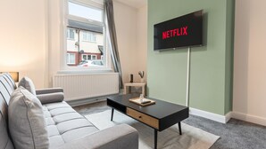 Olohuoneessa on harmaa sohva, musta sohvapöytä, seinälle asennettu televisio, jossa näkyy teksti ”NETFLIX”, ja ikkuna, josta avautuu näkymä ulkona olevaan rakennukseen.