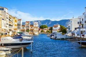 Un port de plaisance où des bateaux sont amarrés le long d’une promenade en bord de mer, entouré de bâtiments dotés de balcons, avec une montagne en arrière-plan.