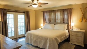 A bedroom with a bed, two bedside lamps, a ceiling fan, a nightstand, and a view through a door. - Cozy lake retreat in Sand Point! Game room with deck and views of Lake Texoma. (Mead)