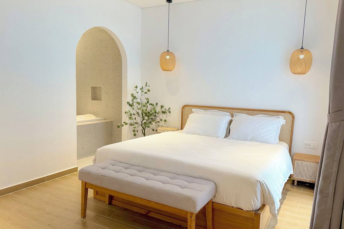 Un dormitorio con una cama de madera, ropa de cama blanca, un banco, una planta y dos lámparas colgantes.