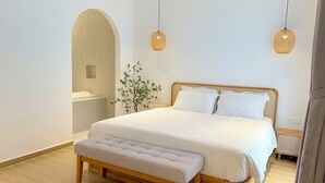 Un dormitorio con una cama de madera, ropa de cama blanca, un banco, una planta y dos lámparas colgantes.