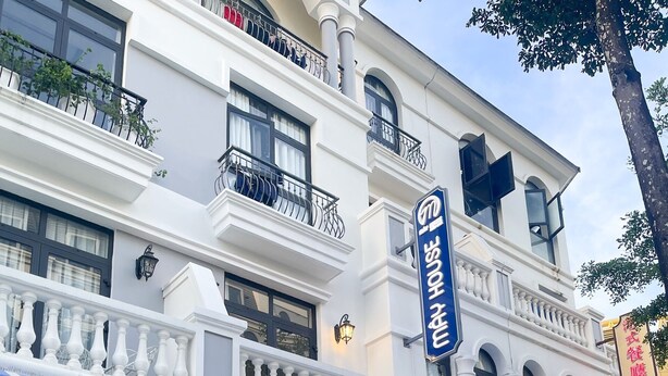 Un hotel con toldo azul y blanco a rayas, un letrero que dice 'MAY HOUSE' y otro que anuncia 'MINI HOTEL COFFEE'.