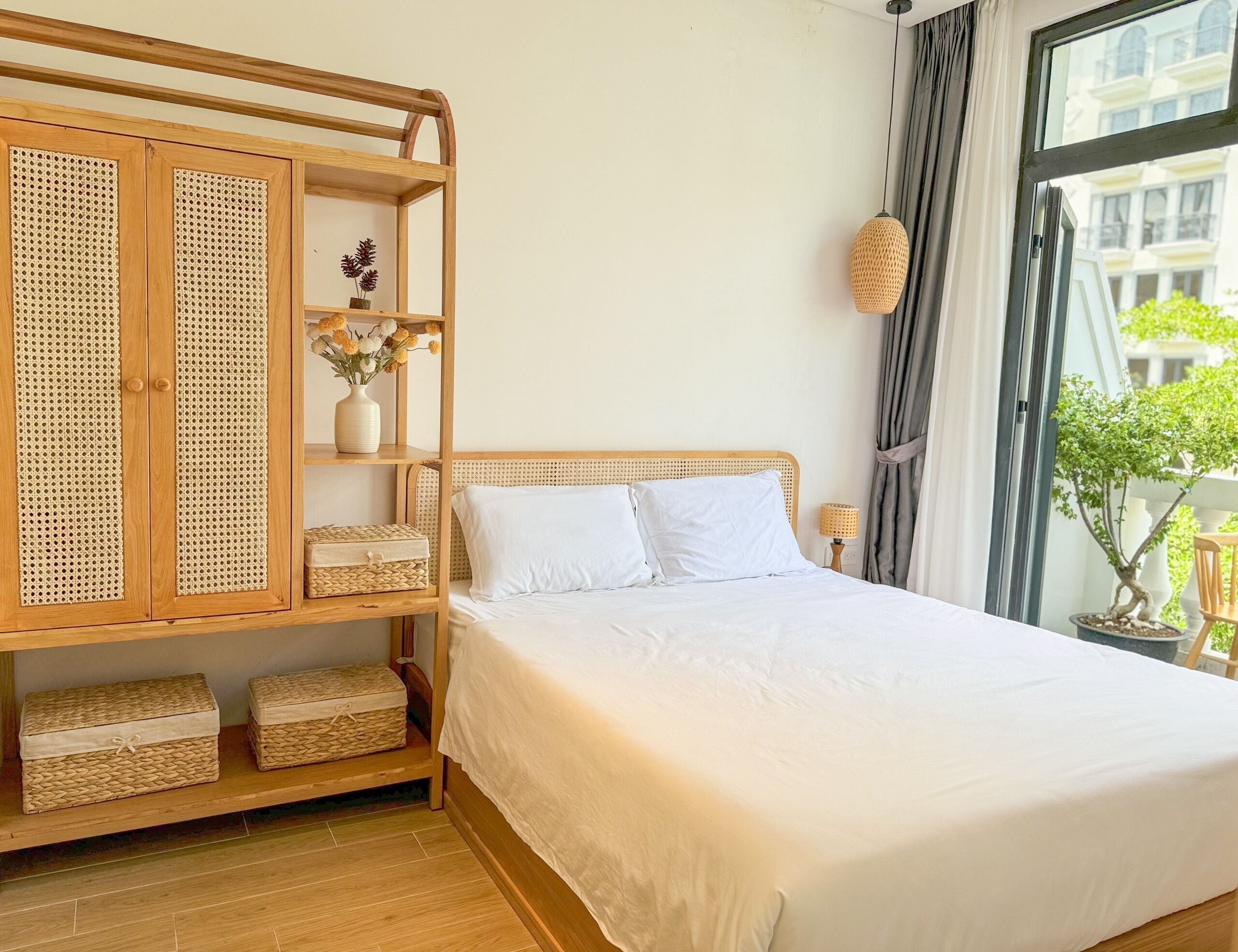 A neatly arranged bedroom with a bed, a wooden wardrobe with woven doors, and a window with a view of buildings and greenery.