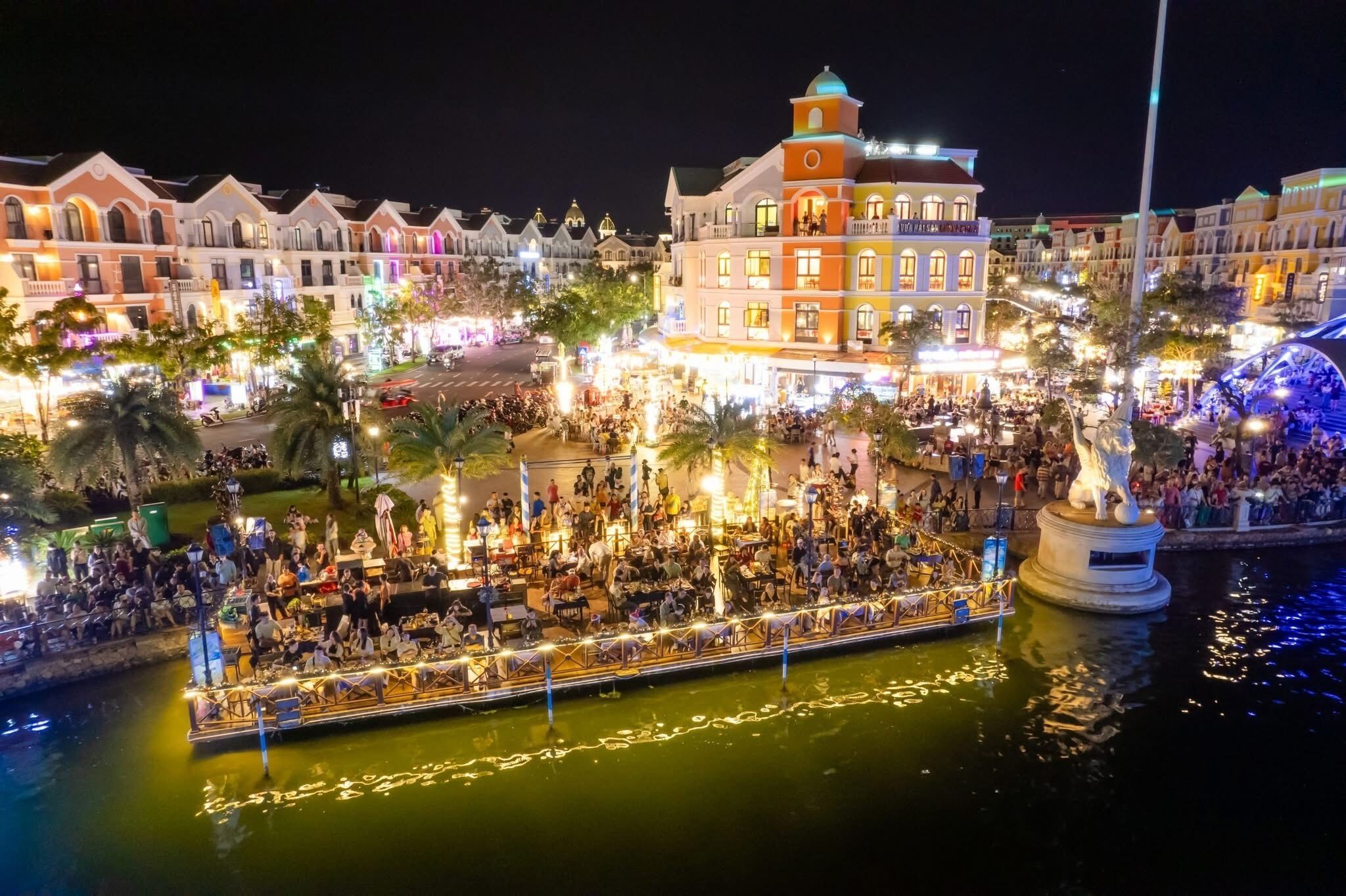Une zone riveraine où un bateau est amarré, entourée de bâtiments aux façades illuminées et d’une statue.