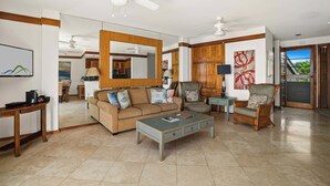 A spacious living room with a large mirror, a sofa, two armchairs, and a coffee table. There is a dining area in the background with a view of the outdoors.