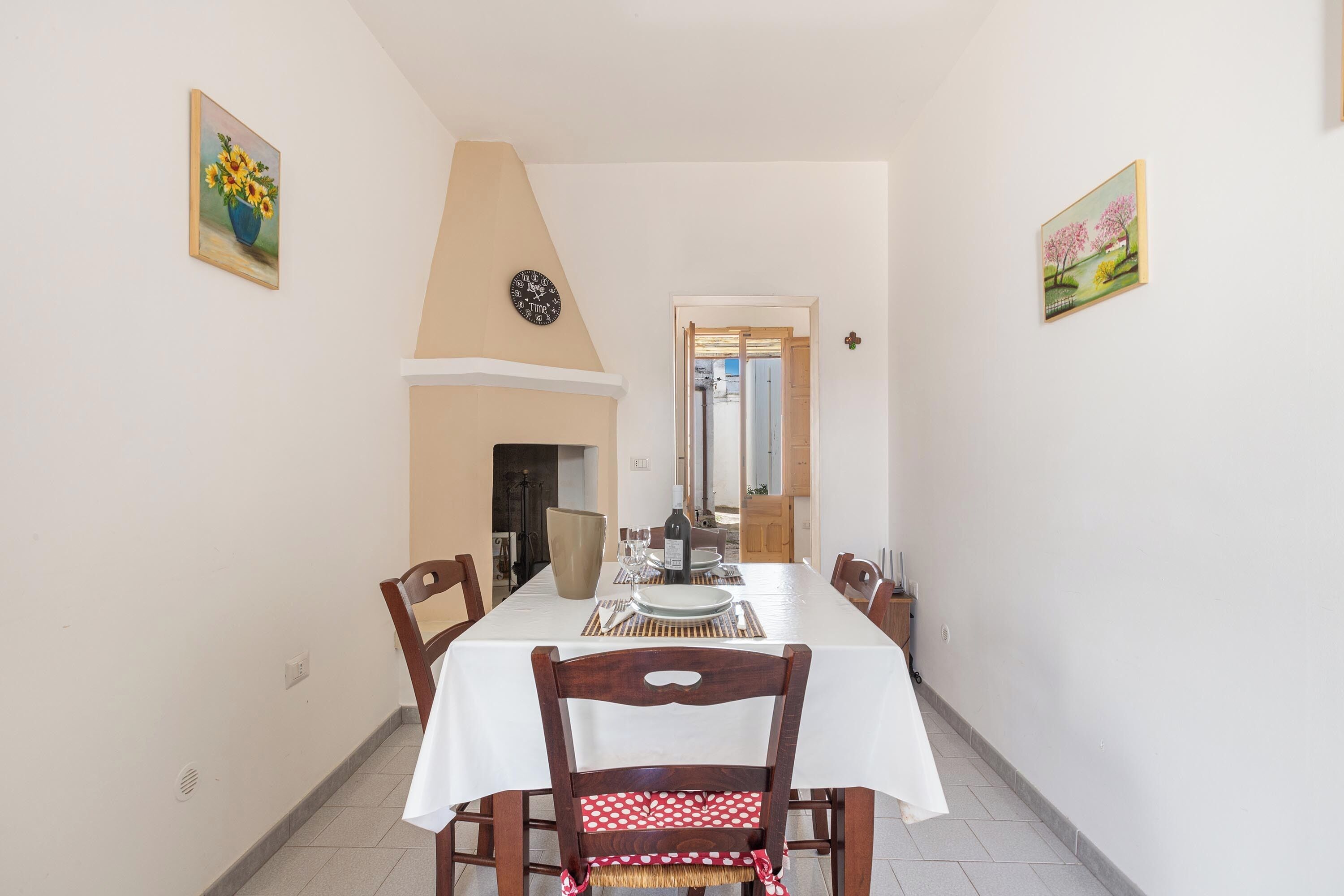 A dining area with a long table, chairs, a clock, and two paintings on the walls.