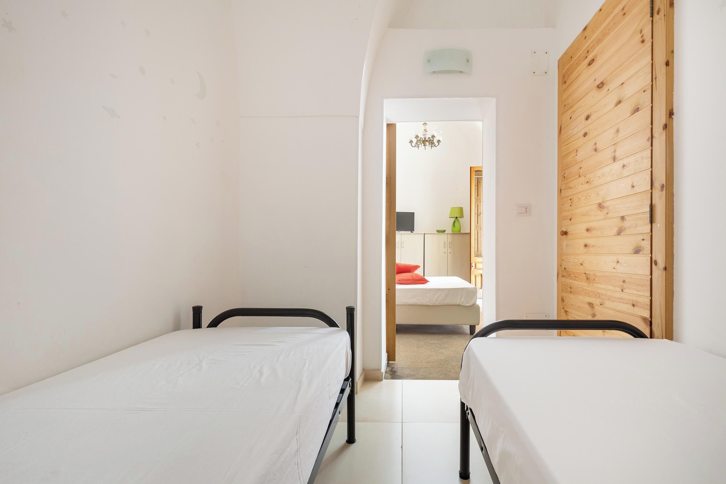 A narrow room with two single beds, a wooden door, and a small hallway leading to another room with a bed and a lamp.