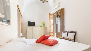 Un dormitorio con una cama, un televisor sobre un mueble, una lámpara verde y una silla de madera con cojines.