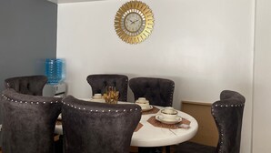 A dining area with a round table, four upholstered chairs, a decorative gold clock, and a blue water dispenser. - Luxurious 2 Bedroom Apartment - Alma by Cytonn (Ruaka)
