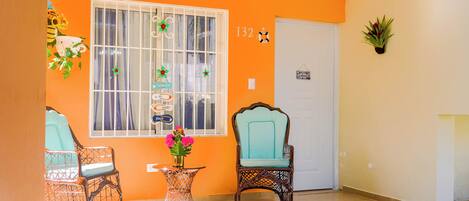 A room with a large window, two wicker chairs, a small table with a vase of flowers, and a door with a sign.