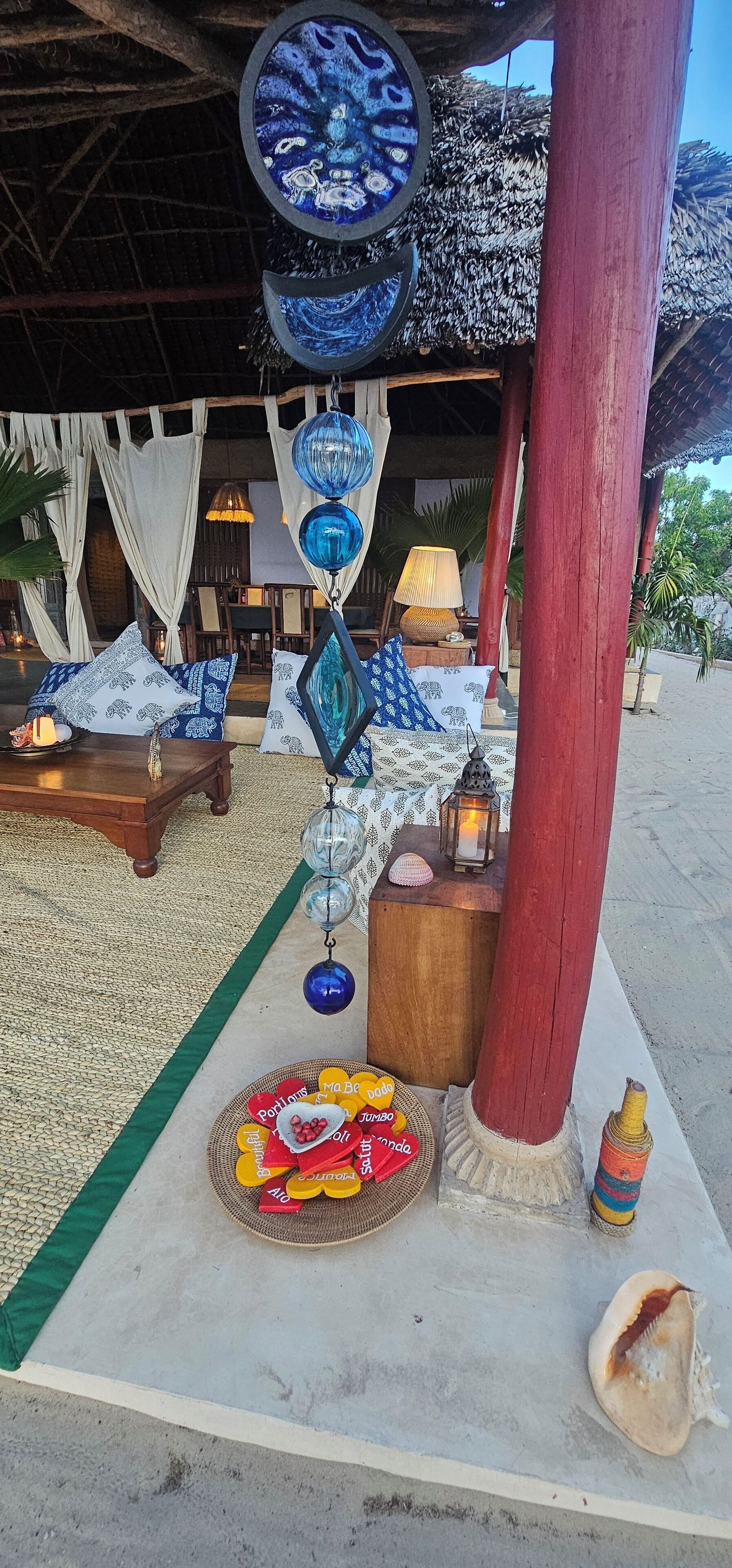 Un espace de détente aménagé sur la plage, avec un toit de chaume, une table en bois agrémentée de coussins et des sphères de verre décoratives suspendues.