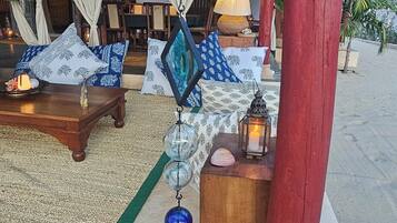 Un espace de détente aménagé sur la plage, avec un toit de chaume, une table en bois agrémentée de coussins et des sphères de verre décoratives suspendues.