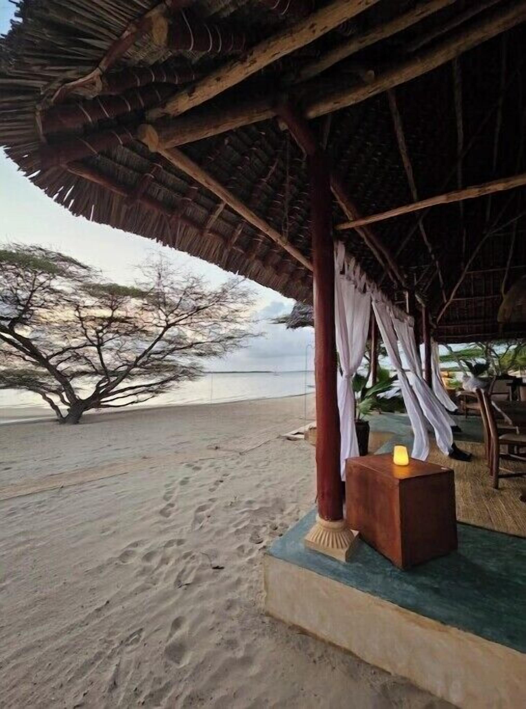 A beachside hut with a thatched roof, a wooden table with a candle, and a tree in the background.
