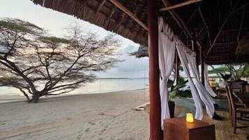 A beachside hut with a thatched roof, a wooden table with a candle, and a tree in the background.
