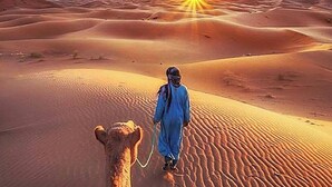 A person in traditional attire walking a camel through a desert landscape with sand dunes and a setting sun. - Merzouga Luxury Camp (Taouz)