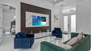 A modern living room with a large flat-screen TV, two blue armchairs, a green sofa, and a glass coffee table. - Modern Vacation Home Fully Fences Near Airport (Lehigh Acres)