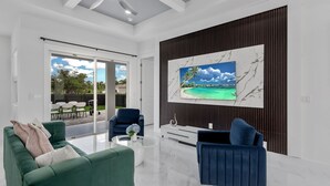 A modern living room with a green sofa, a blue armchair, and a coffee table. There is a large TV mounted on a wall with a tropical beach scene. - Modern Vacation Home Fully Fences Near Airport (Lehigh Acres)