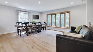 Amplia sala de estar con una mesa de comedor y sillas, un sofá de cuero negro, un televisor de pantalla plana montado en la pared y grandes ventanas con vistas a árboles.
