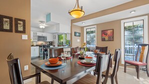 A dining area with a wooden table set for six, surrounded by chairs. A kitchen is visible in the background, and there is a large window with a view of greenery outside. - Spacious 5-Bedroom House in Rockville Fast WiFi, Gaming Area & Fenced Yard (Rockville)