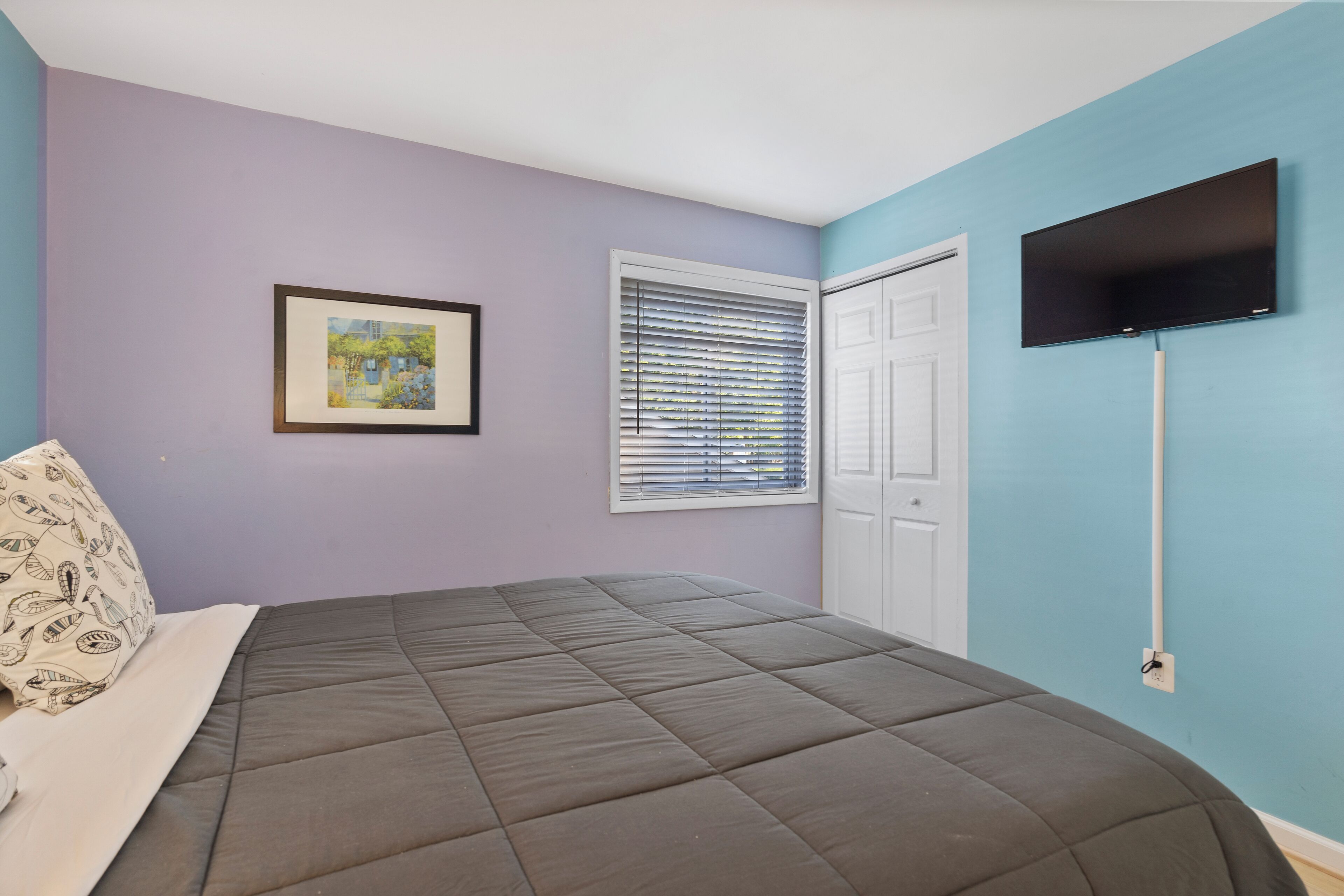 A bedroom with a bed, a window with blinds, a door, and a TV mounted on the wall.