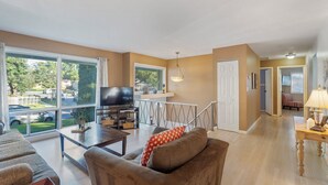A spacious living room with a large TV, comfortable seating, and a balcony with a view of the street. - Spacious 5-Bedroom House in Rockville Fast WiFi, Gaming Area & Fenced Yard (Rockville)
