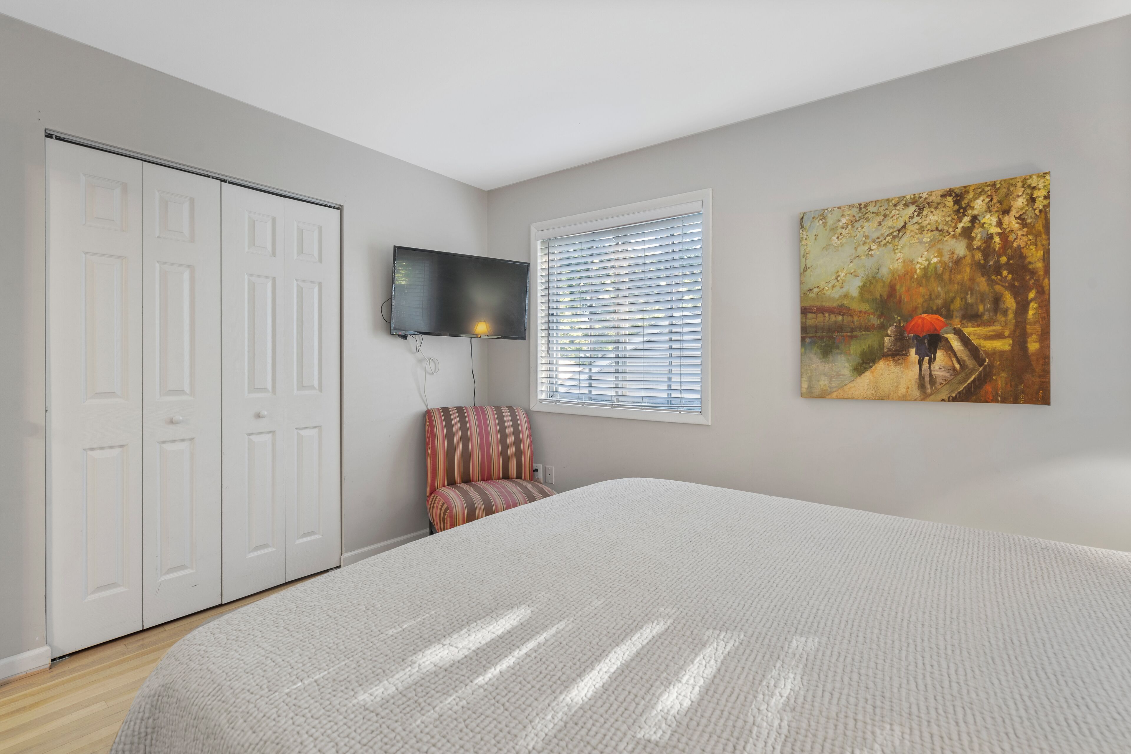 A bedroom with a large bed, a TV mounted on the wall, a window with blinds, and a painting on the wall.