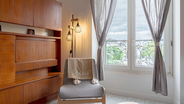 Une pièce avec un meuble en bois, une chaise recouverte d’une couverture ornée du mot « Charm », un luminaire fixé au mur et une fenêtre avec des rideaux.