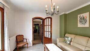 A room with a wooden chair, a sofa, a table, a chandelier, and a door leading to another room. - Vert Olive - Maison proche Sélestat, Colmar (Rombach-le-Franc)