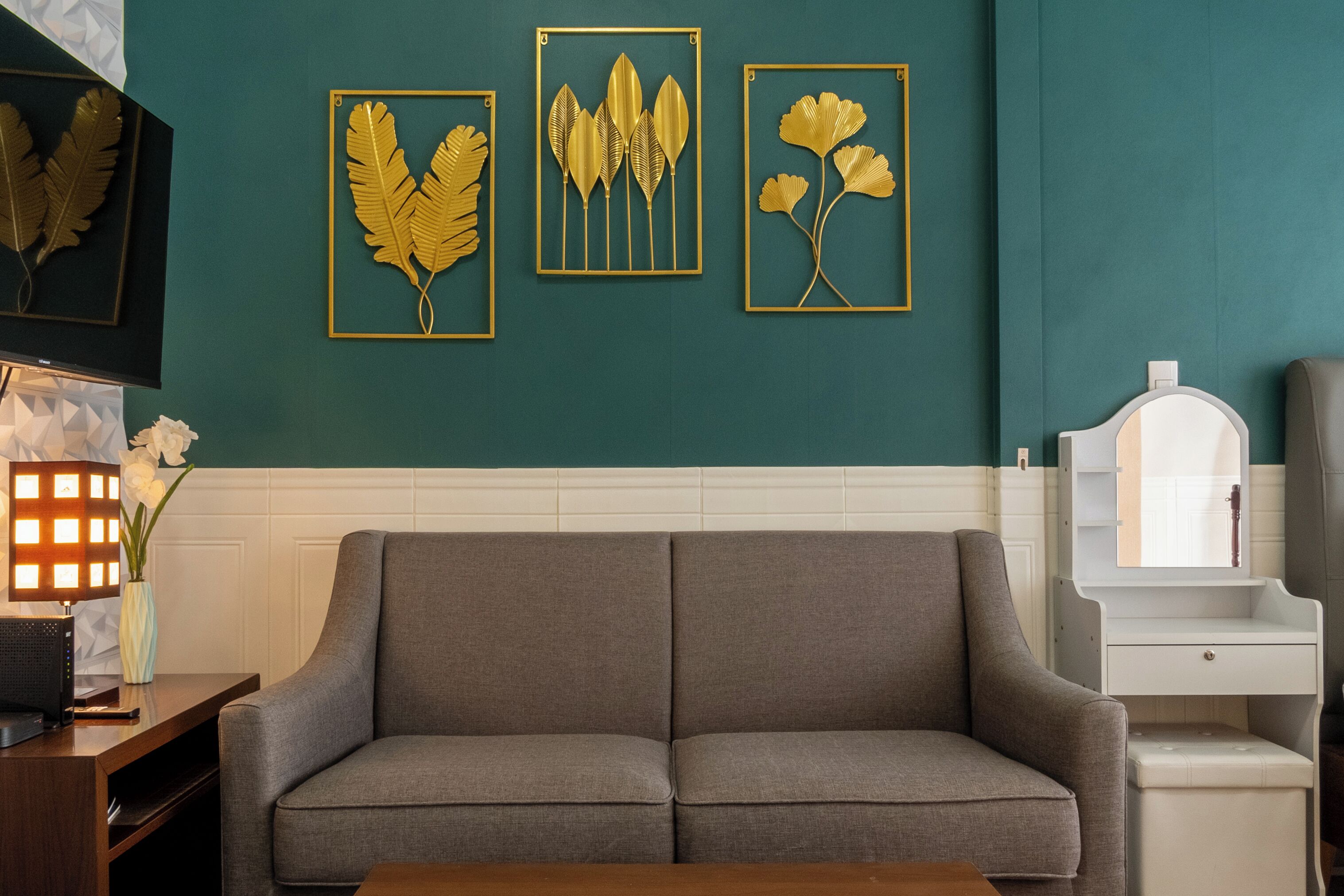 Una habitación con un sofá gris, una mesita de madera con una lámpara, y tres obras de arte con hojas doradas enmarcadas sobre paredes verde azulado.