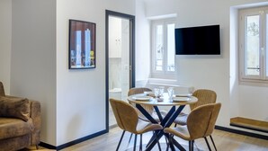 A modern living room with a round dining table, comfortable chairs, a flat-screen TV mounted on the wall, and a large window providing natural light. - Le Turbiasque - apt climatisé - 500m de la plage (La Turbie)