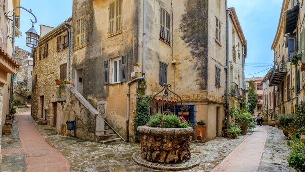 A narrow cobblestone street flanked by historic buildings with stone facades, a central stone fountain, and potted plants. - Le Turbiasque - apt climatisé - 500m de la plage (La Turbie)