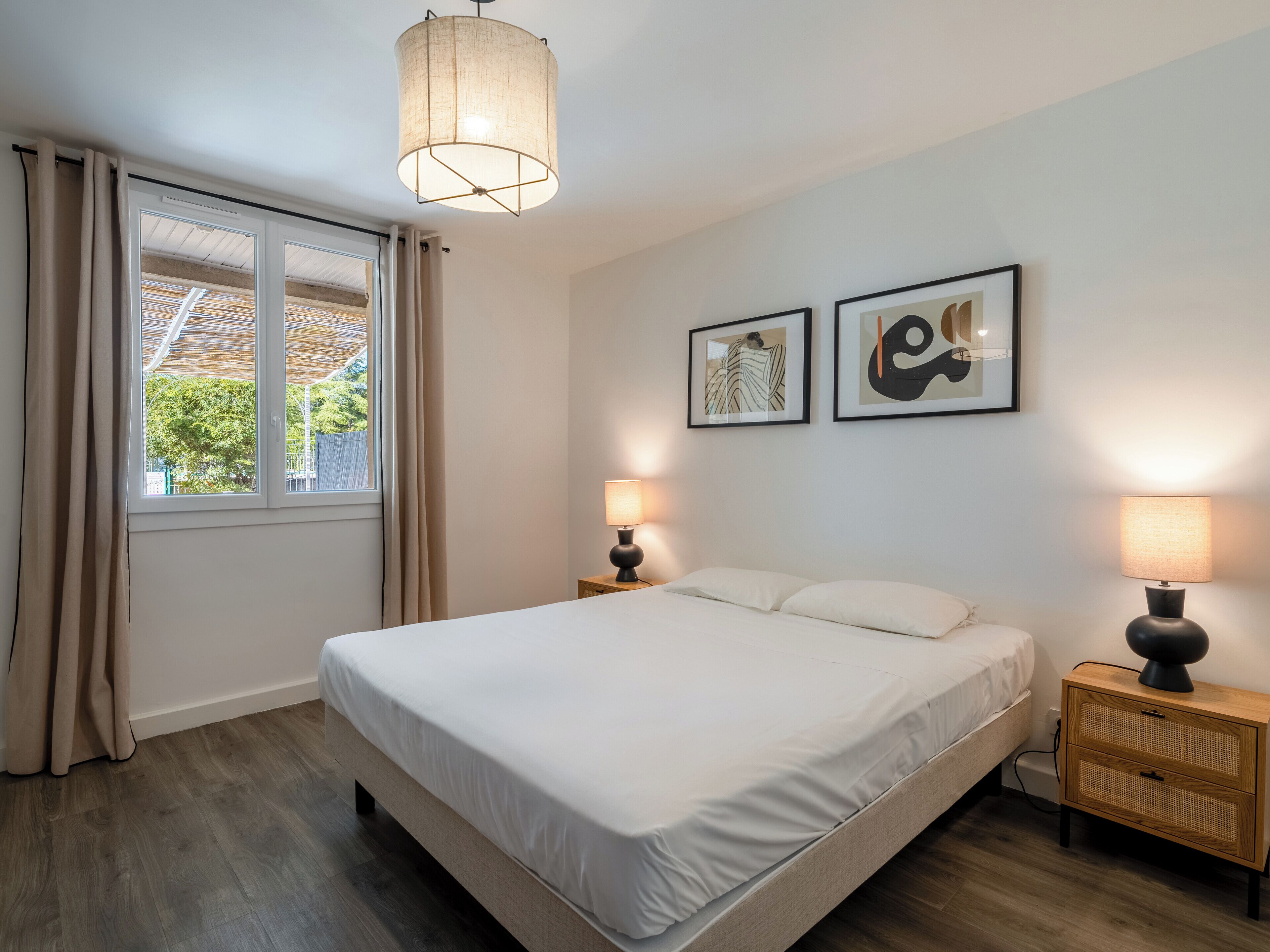 Een slaapkamer met een bed, twee bedlampen, een raam met gordijnen en twee ingelijste kunstwerken aan de muur.