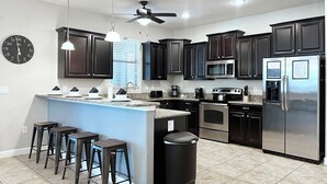 Une cuisine moderne avec des meubles de couleur sombre, des appareils en acier inoxydable, un îlot central doté de tabourets et un ventilateur de plafond.