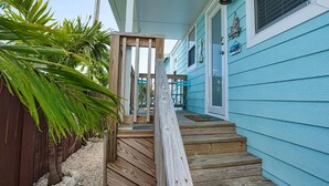 Un escalier en bois mène à une maison bleue ornée d’une pancarte « Bienvenue », ainsi qu’à une clôture en bois et à un feuillage verdoyant.