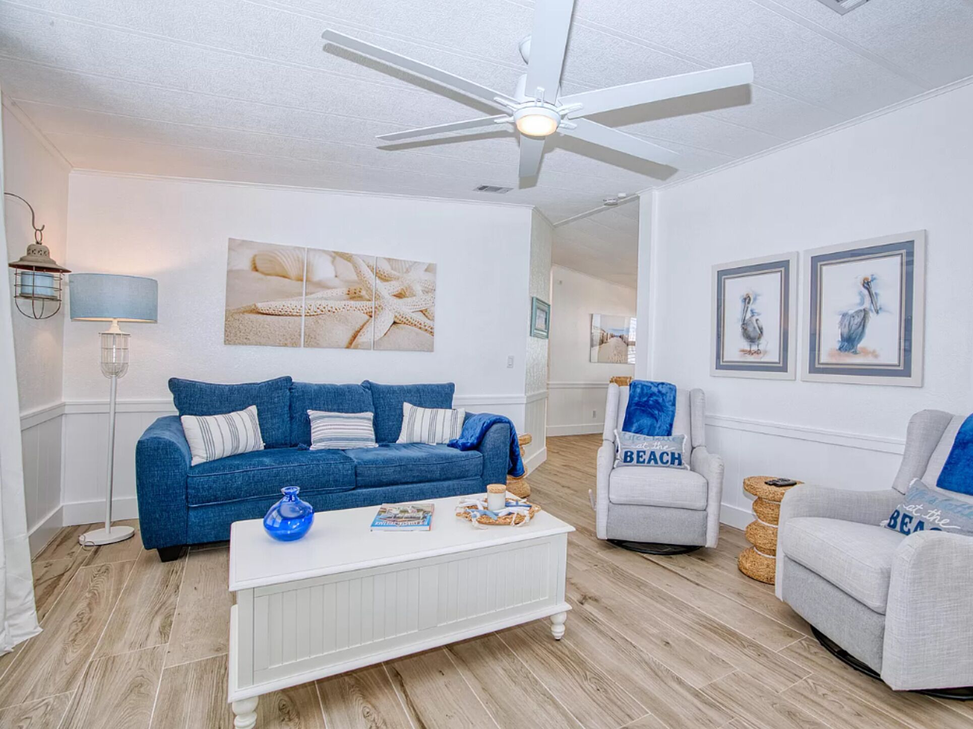 A living room with a blue sofa, a white coffee table, and a grey armchair. There is a ceiling fan, a lamp, and artwork on the walls.