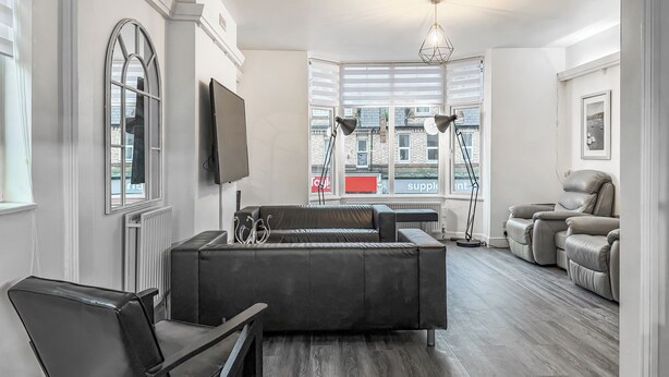 A modern living room with a sectional sofa, a recliner, a TV mounted on the wall, and a large window with blinds. - Discover a spacious holiday home in Paignton, Devon, accommodating ten guests. (Paignton)