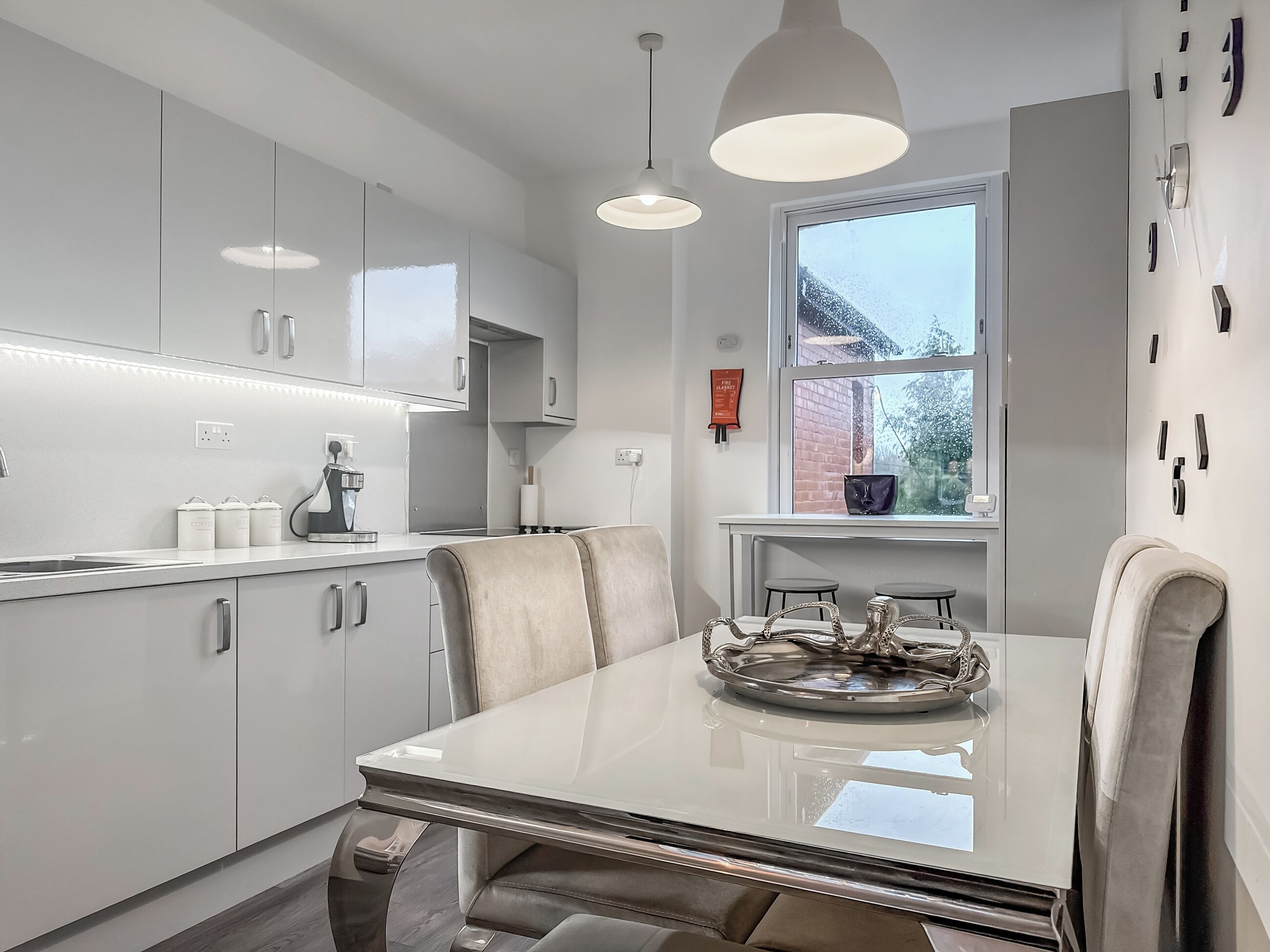 Une cuisine moderne avec des meubles blancs, une table à manger avec un plateau en verre et des luminaires suspendus.