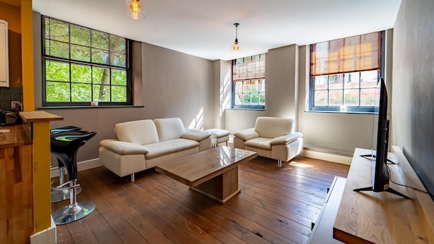 A modern living room with a white sofa, a coffee table, and a TV on a wooden stand. There are large windows with blinds, and a wooden floor. - Stunning Modern City Centre Apartment Retreat (Nottingham)