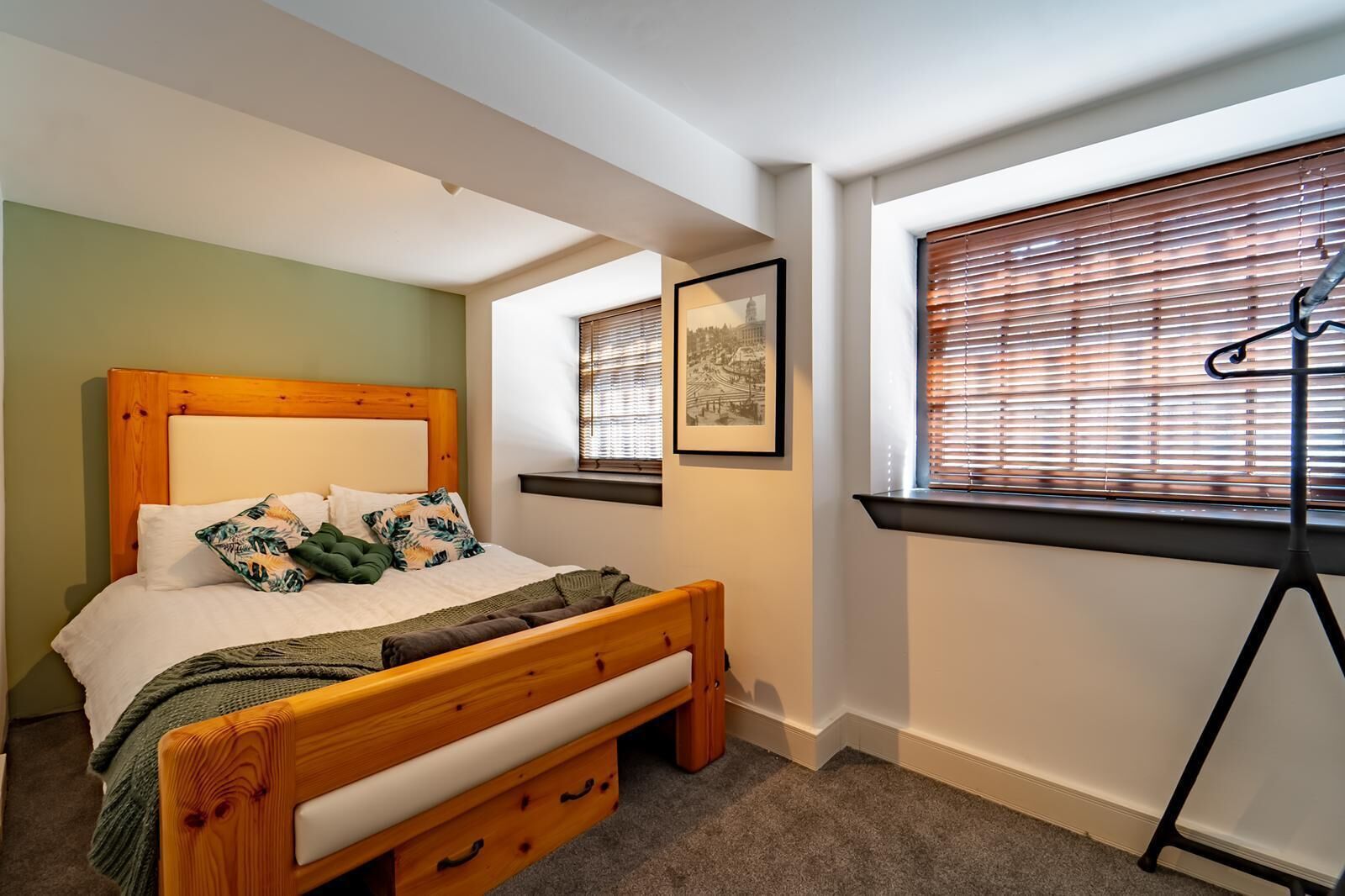 Une chambre avec un lit en bois, une fenêtre avec des stores, un tableau encadré au mur et une petite table avec une chaise.