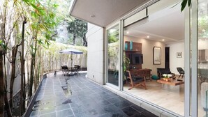Un patio moderno al aire libre con piso de baldosas negras curvas, una mesa y sillas de madera, y una puerta de vidrio que da a un espacio interior con un sofá y un televisor.