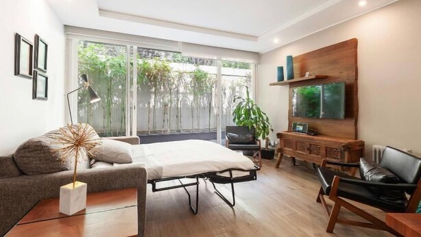 Una sala moderna con sofá, mesa de centro, cama, televisor y un ventanal con vistas al verdor.