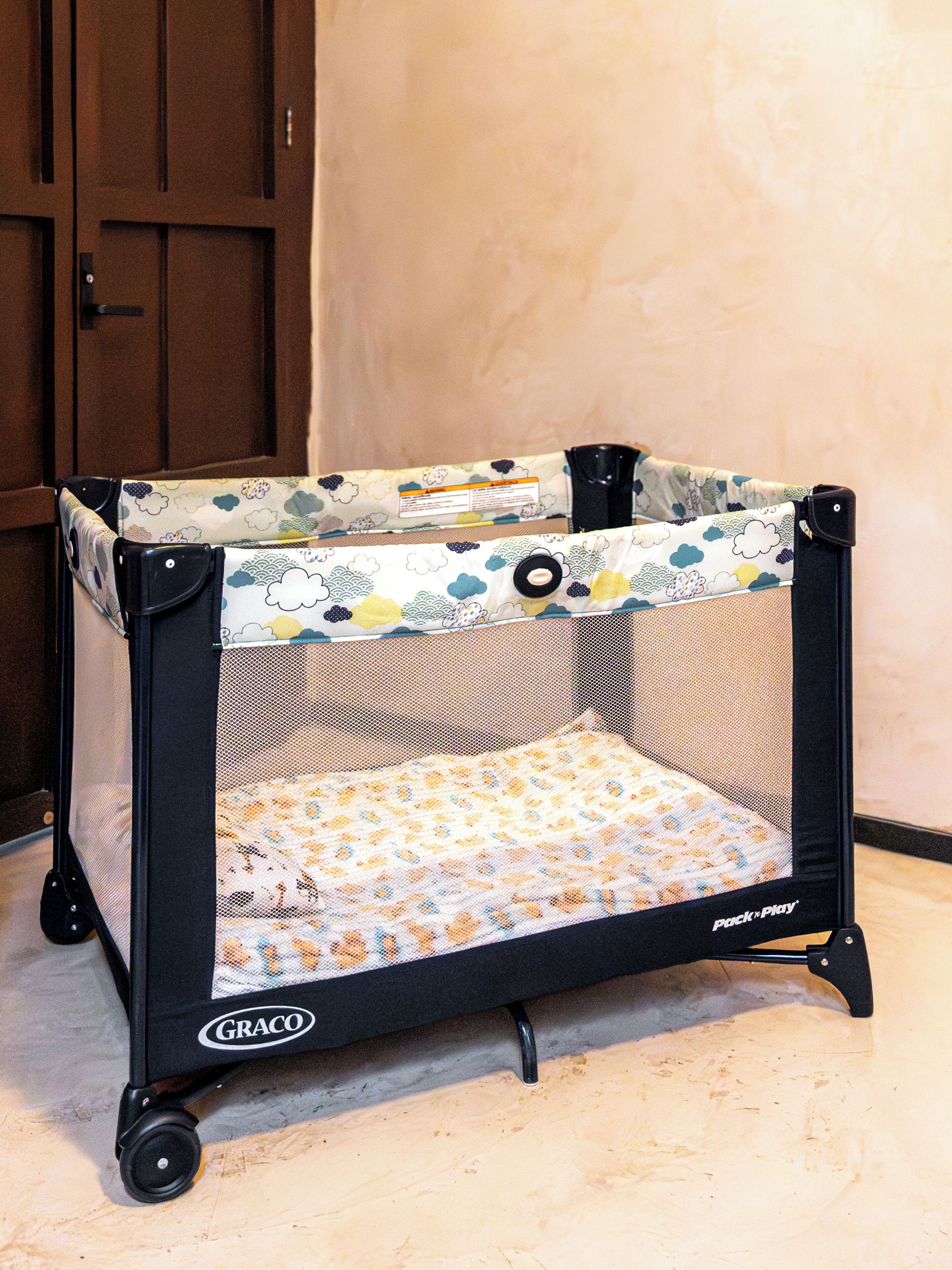 Ein tragbares Reisebett mit Maschennetzoberteil und einem gemusterten Kissen, mit dem Aufdruck 'Pack n Play' von Graco, auf Rollen.