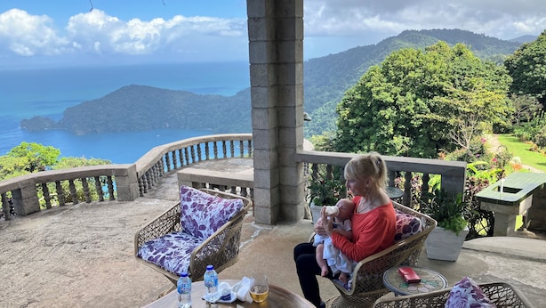 A balcony with wicker furniture, a table with water bottles, and a view of the ocean and hills.