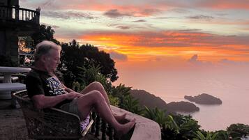 A man sitting on a bench overlooking a sunset with a view of the sea and distant islands.